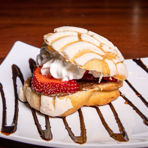 A concha bread dessert filled with whipped cream, strawberries, and chocolate drizzle on a white plate.