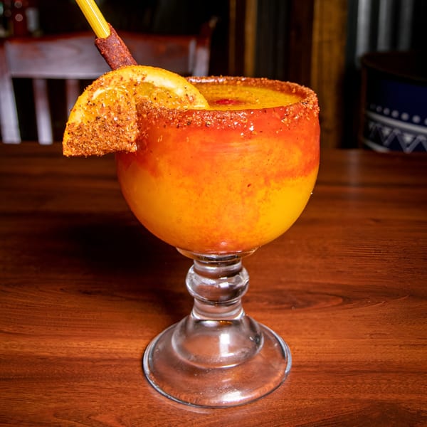 A vibrant orange and yellow drink in a goblet, garnished with a chili-dusted orange slice and straw.