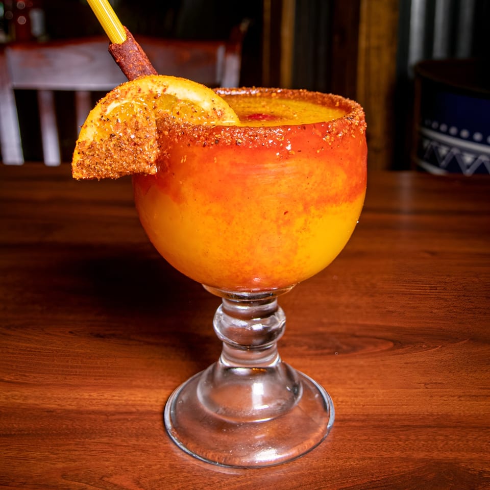 A vibrant orange and yellow drink in a goblet, garnished with a chili-dusted orange slice and straw.