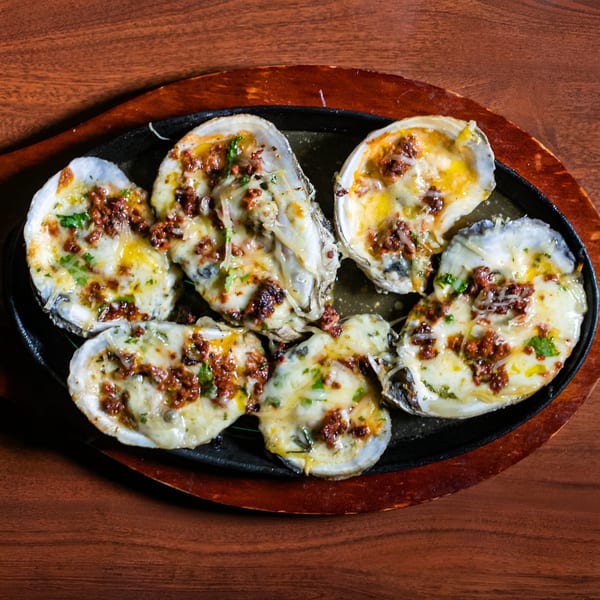 Six baked oysters topped with melted cheese, chorizo, and herbs, served on a sizzling platter.