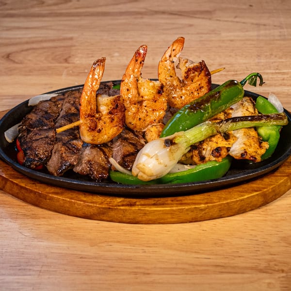 Sizzling platter of grilled shrimp skewers, steak, chicken, green peppers, and onions on wood.